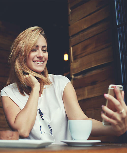 A photo of a young woman holding a mobile phone and smiling. This image is meant to communicate the nature of how we'll work together as a coach and client to get you pleasing results.